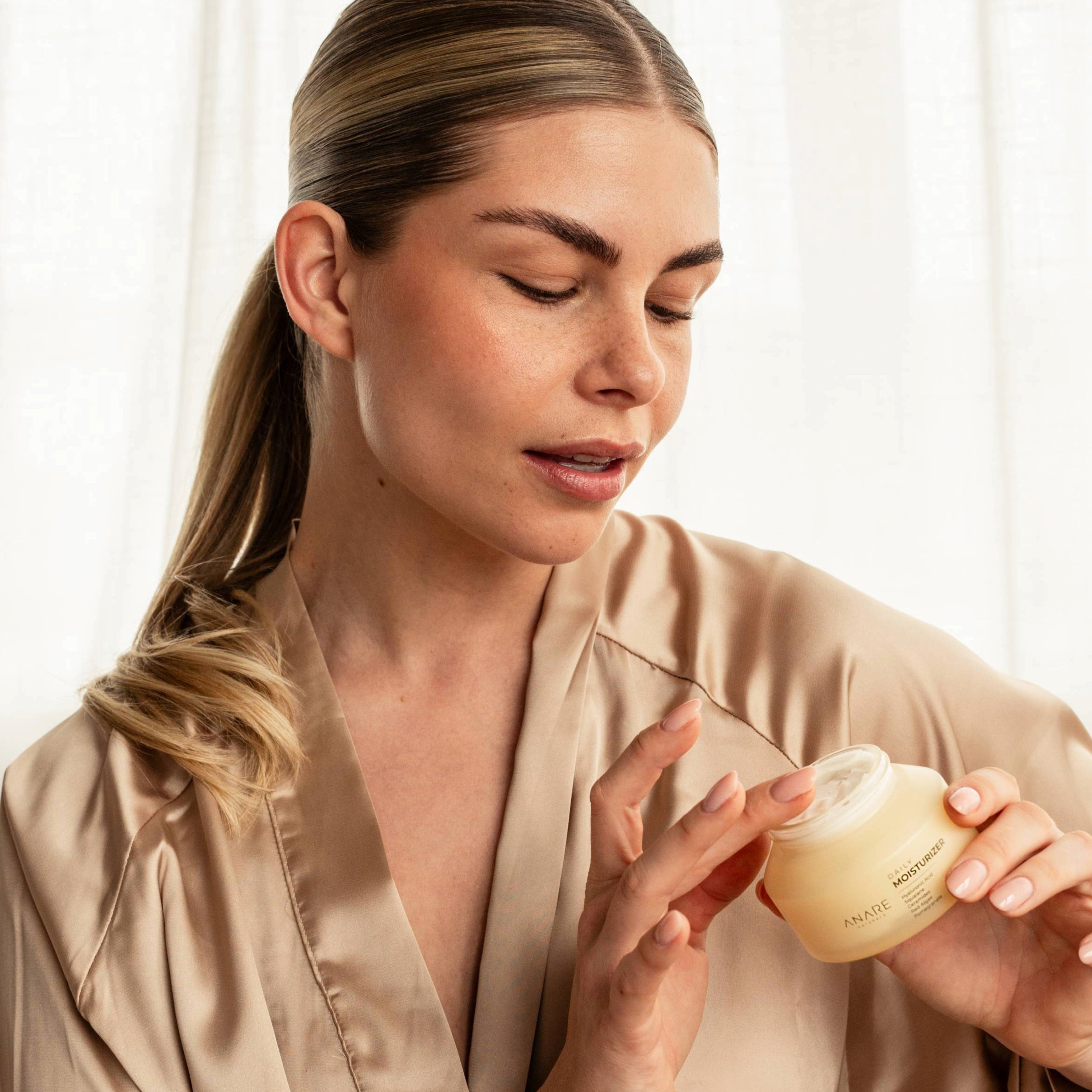 Blonde model with brown eyes demonstrating Anare Naturals Daily Moisturizer while sitting on a couch in a morning bathrobe.