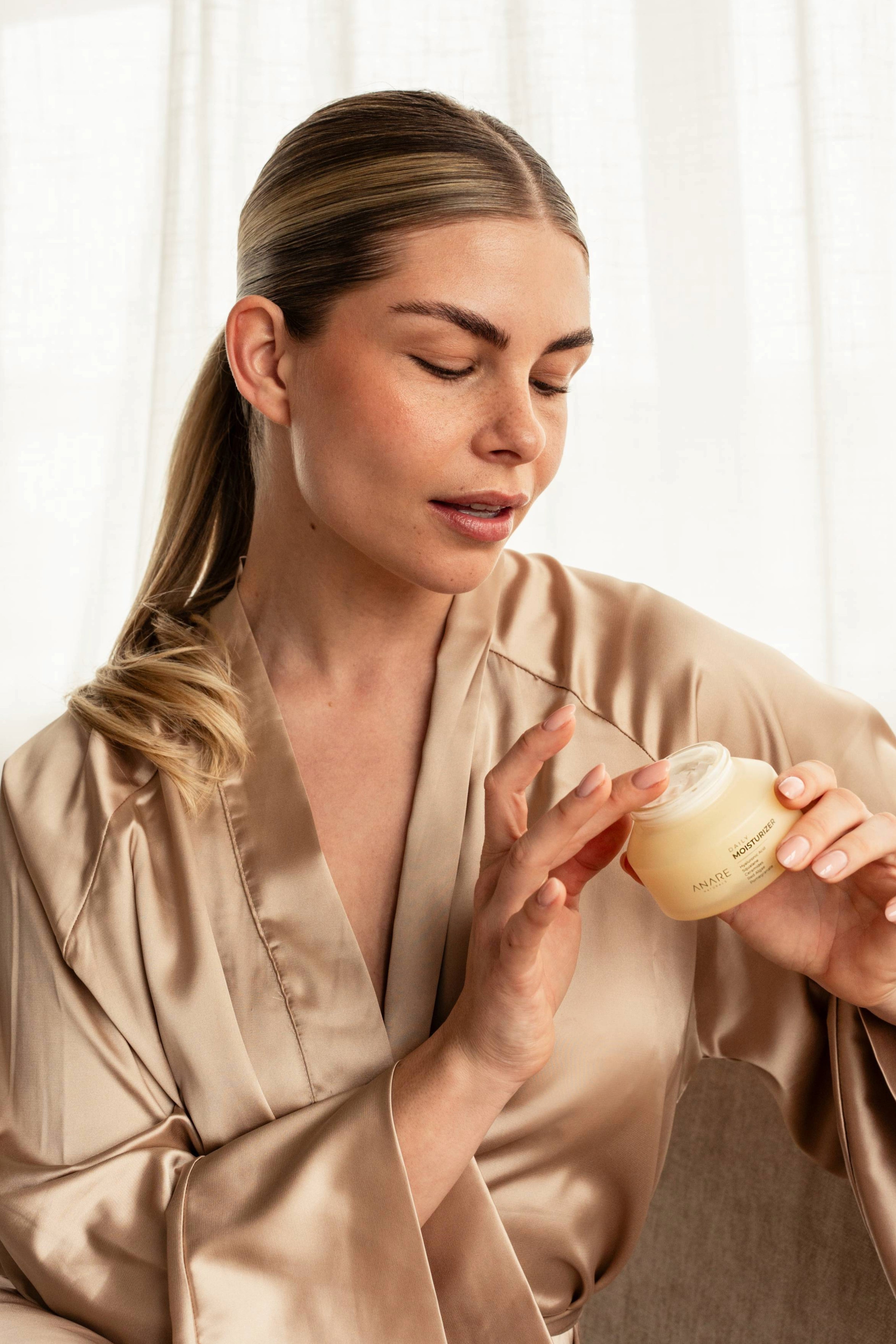Blonde model with brown eyes demonstrating Anare Naturals Daily Moisturizer while sitting on a couch in a morning bathrobe.