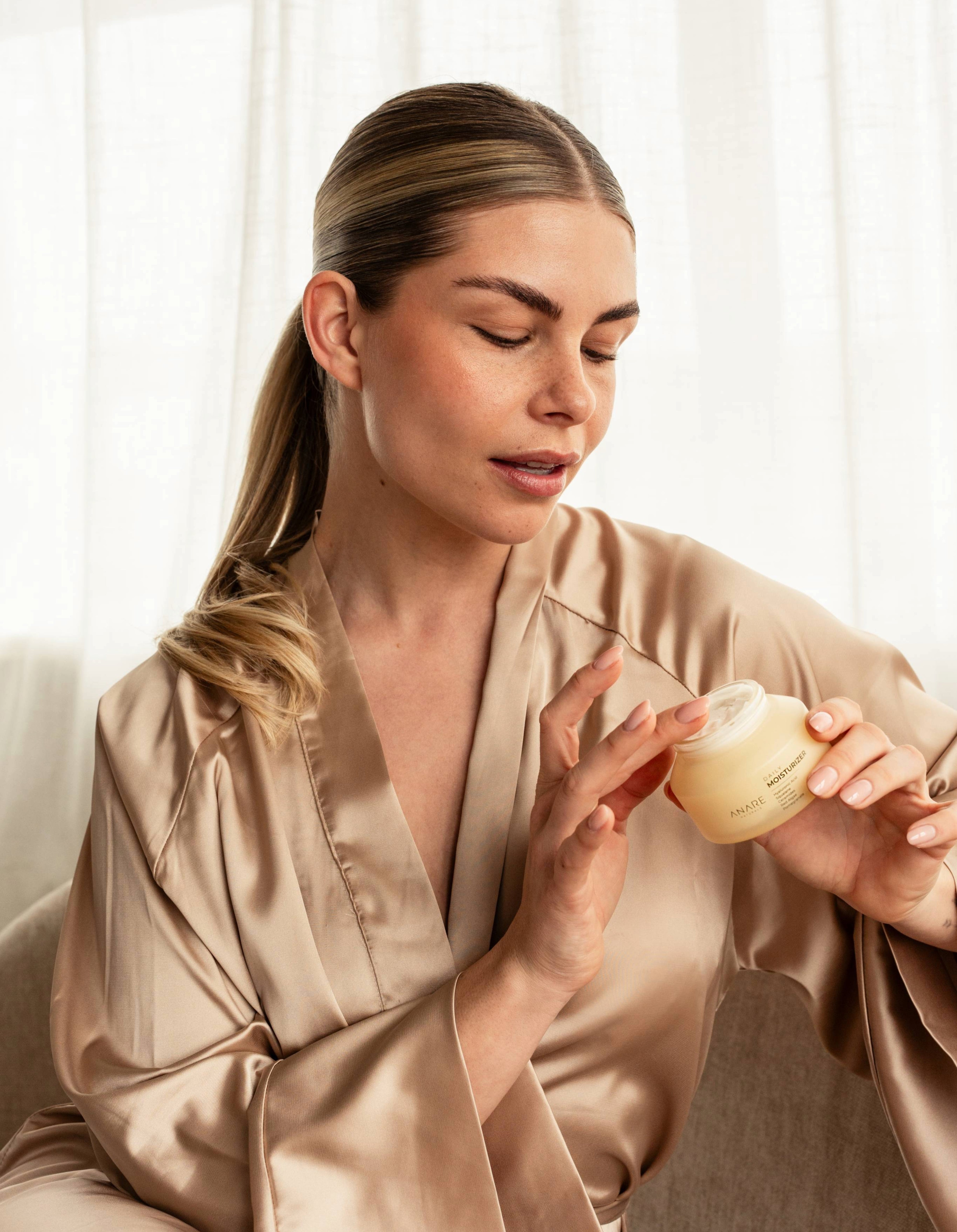 Blonde model with brown eyes demonstrating Anare Naturals Daily Moisturizer while sitting on a couch in a morning bathrobe.
